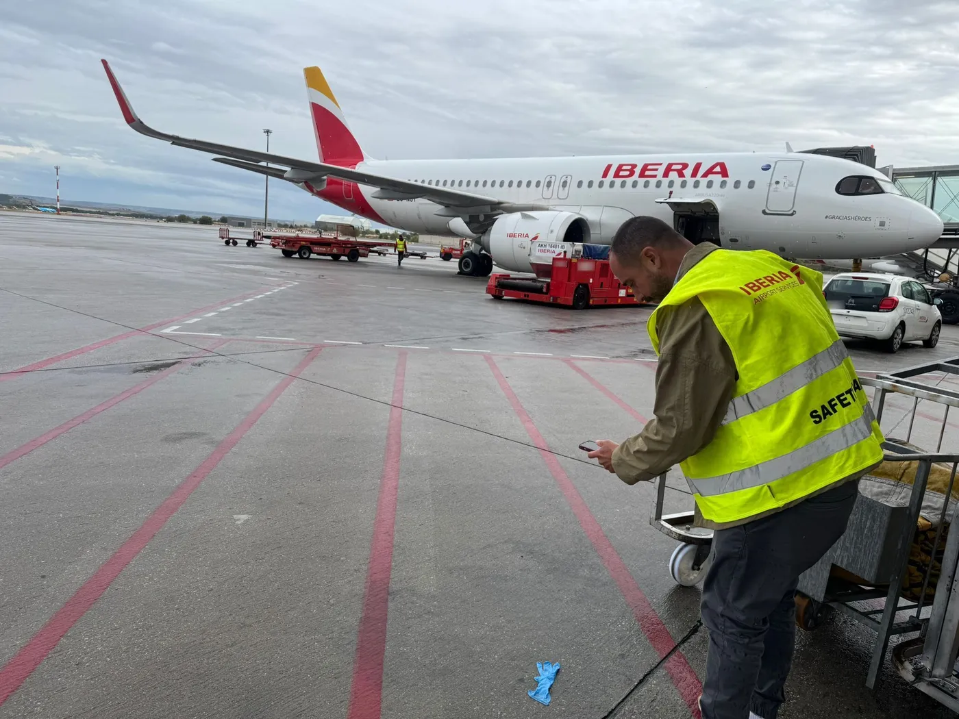 A ground handler photographs debris on a live apron with a phone; behind them, turnaround continues.
